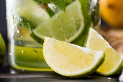 A close-up of a fresh lime wedge in the foreground, with a chilled glass of a green mojito drink containing more lime slices and mint leaves in the background.