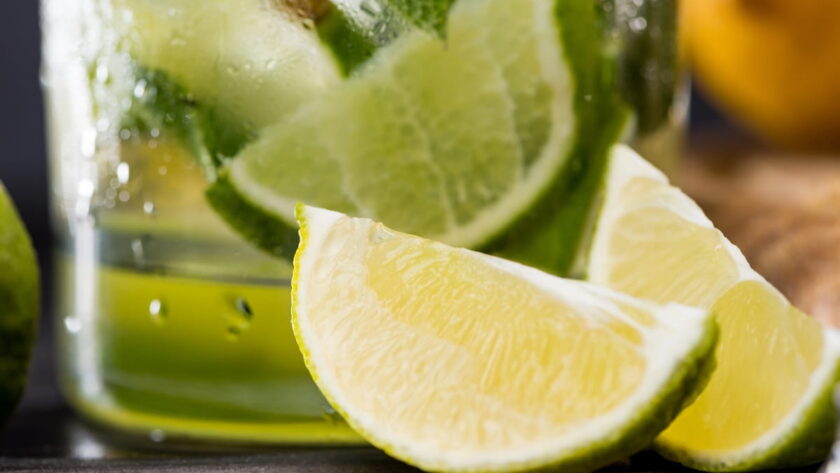 A close-up of a fresh lime wedge in the foreground, with a chilled glass of a green mojito drink containing more lime slices and mint leaves in the background.