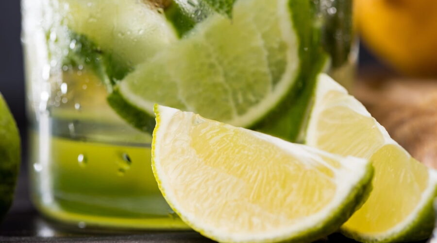 A close-up of a fresh lime wedge in the foreground, with a chilled glass of a green mojito drink containing more lime slices and mint leaves in the background.