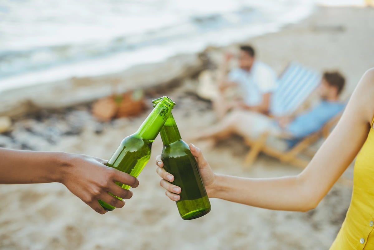 Two hands clink green glass bottles together in a toast on a sandy beach. In the blurred background, a group of friends relax in lounge chairs near the ocean waves, perfectly capturing the social and carefree spirit of a beach lime.