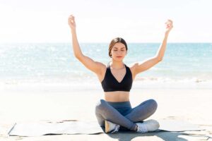 Woman mediating with arms raised on yoga mat at beach during summer vacation.