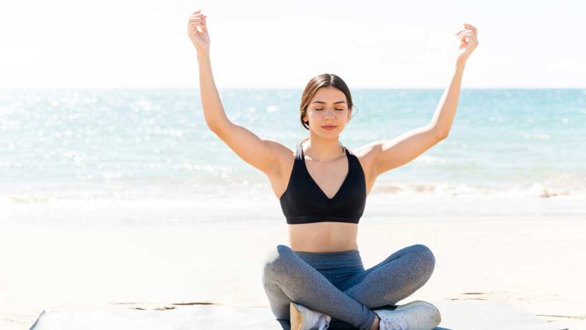 Woman mediating with arms raised on yoga mat at beach during summer vacation.