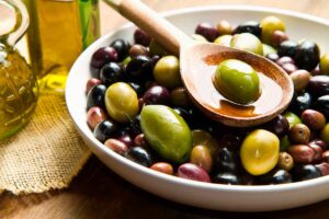 Bowl of mixed Italian olives in oil with a wooden spoon, next to a glass bottle of olive oil on a rustic table.