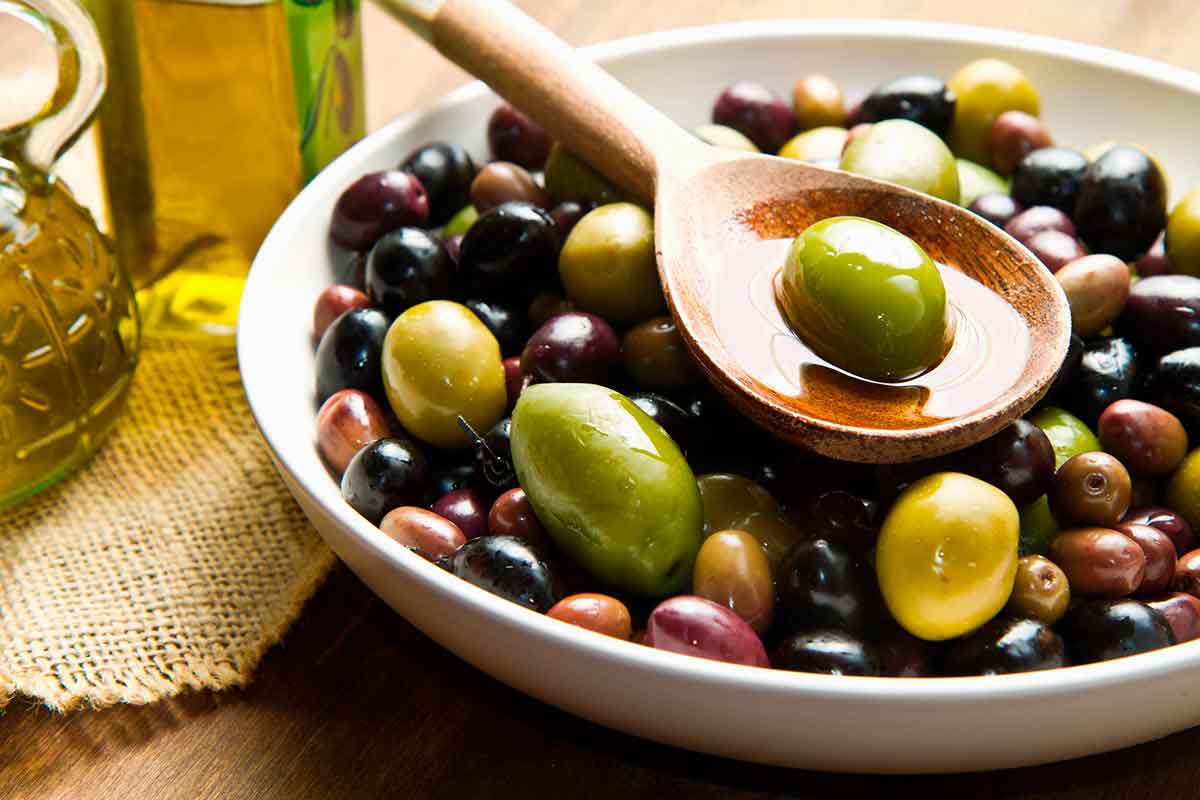 Bowl of mixed Italian olives in oil with a wooden spoon, next to a glass bottle of olive oil on a rustic table.
