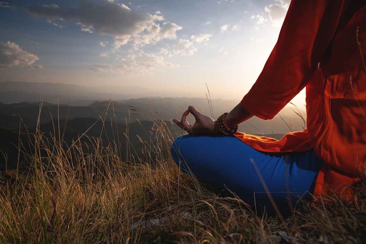 Close-up of a woman and female hands sitting in a lotus position, yoga outdoors in the grass at sunset against the backdrop of a mountain range and clouds, meditation.