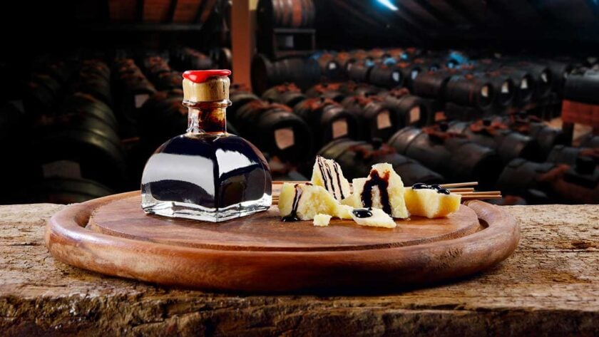 Small glass bottle of dark balsamic vinegar with a red wax seal, placed on a wooden serving board alongside chunks of cheese drizzled with balsamic vinegar. In the background, rows of aging barrels are visible inside a traditional balsamic vinegar cellar.