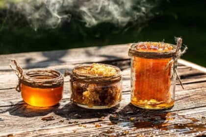 Three rustic glass jars of various sizes filled with golden raw honey and fresh honeycomb, sitting on a weathered wooden table with smoke drifting in the background.