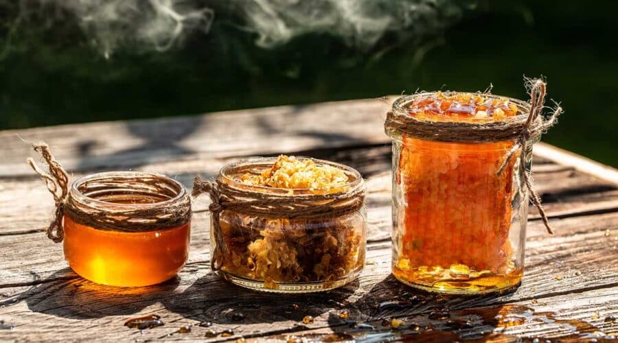 Three rustic glass jars of various sizes filled with golden raw honey and fresh honeycomb, sitting on a weathered wooden table with smoke drifting in the background.