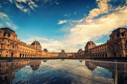 Wide-angle view of the Louvre Museum courtyard in Paris under a dramatic blue and white sky, featuring the historic palace architecture and the central glass pyramid perfectly reflected in a glass surface below.
