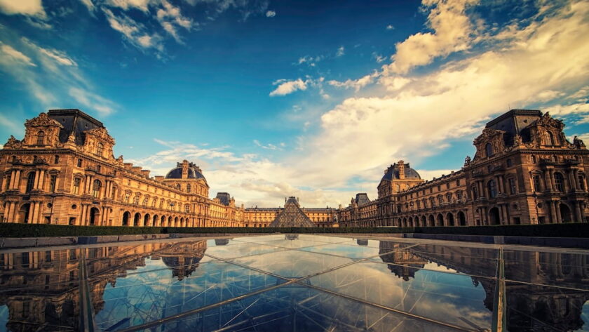 Wide-angle view of the Louvre Museum courtyard in Paris under a dramatic blue and white sky, featuring the historic palace architecture and the central glass pyramid perfectly reflected in a glass surface below.
