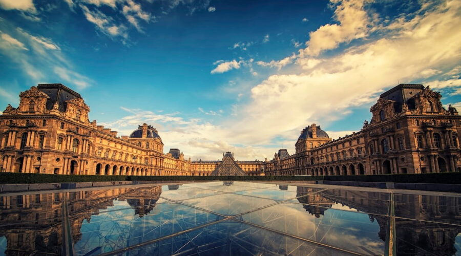 Wide-angle view of the Louvre Museum courtyard in Paris under a dramatic blue and white sky, featuring the historic palace architecture and the central glass pyramid perfectly reflected in a glass surface below.