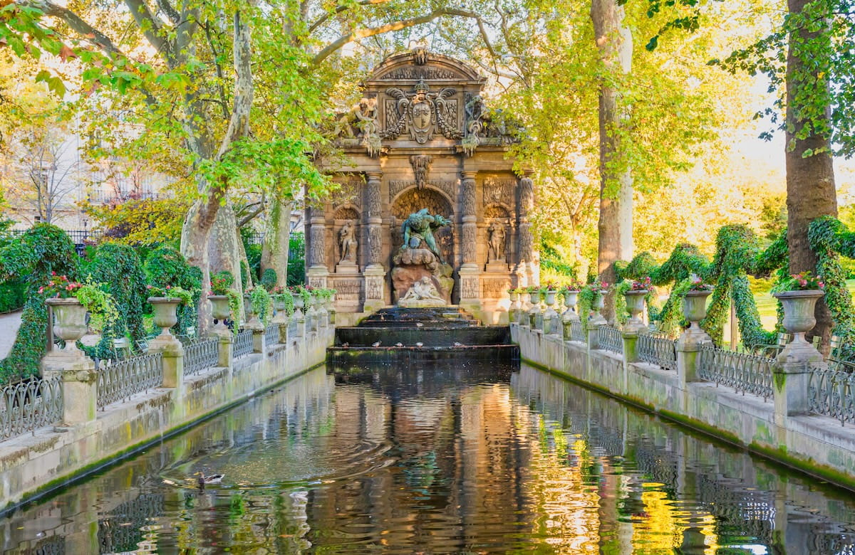 The historic Medici Fountain in the Luxembourg Garden in Paris, featuring an ornate stone monument with classical statues at the end of a long, narrow reflecting pool lined with ivy-draped railings and stone flower urns.