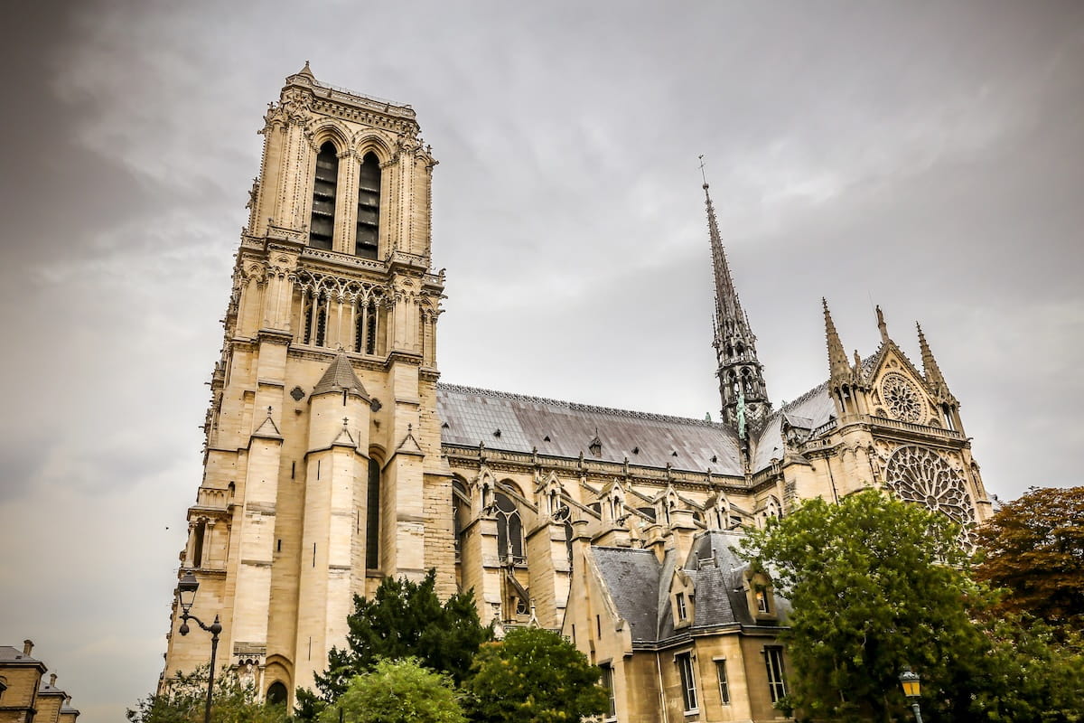Angled side view of the Notre-Dame Cathedral in Paris under an overcast, cloudy sky, showcasing its Gothic architecture, soaring stone towers, central spire, and intricate rose window.