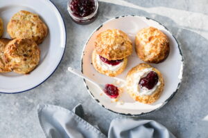 A flat-lay, top-down view of a cream tea spread on a grey stone surface. On a white enamel plate with a distressed black rim, there are almond-topped scones, including one split open and layered with clotted cream and dark red jam, and another open-faced half topped with cream and jam. A small white spoon with a bit of jam sits on the plate alongside a blue-rimmed plate of extra scones, an open jar of dark berry jam, and a folded grey cloth napkin.
