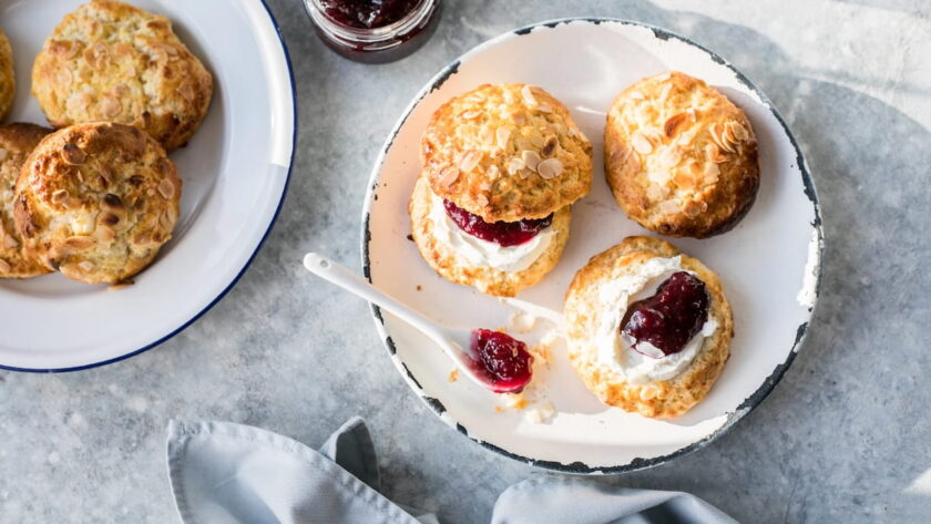 A flat-lay, top-down view of a cream tea spread on a grey stone surface. On a white enamel plate with a distressed black rim, there are almond-topped scones, including one split open and layered with clotted cream and dark red jam, and another open-faced half topped with cream and jam. A small white spoon with a bit of jam sits on the plate alongside a blue-rimmed plate of extra scones, an open jar of dark berry jam, and a folded grey cloth napkin.