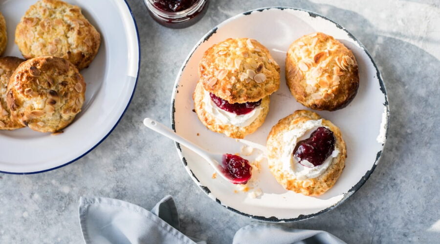 A flat-lay, top-down view of a cream tea spread on a grey stone surface. On a white enamel plate with a distressed black rim, there are almond-topped scones, including one split open and layered with clotted cream and dark red jam, and another open-faced half topped with cream and jam. A small white spoon with a bit of jam sits on the plate alongside a blue-rimmed plate of extra scones, an open jar of dark berry jam, and a folded grey cloth napkin.