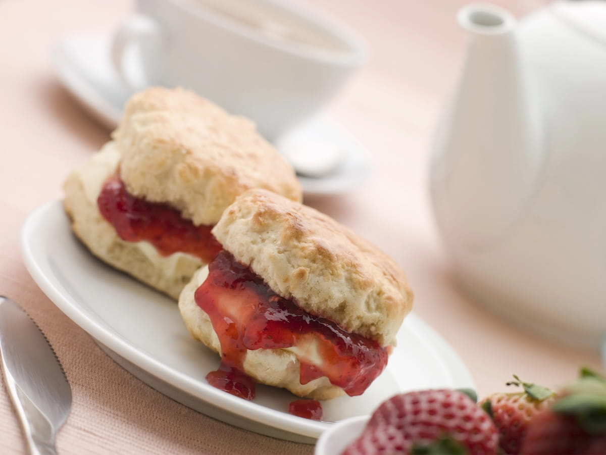 A bright, soft-focus shot of a traditional cream tea setting. Two golden-brown scones, split open and layered with thick clotted cream and dripping red strawberry jam, rest on a white ceramic plate. In the blurred background, a white teapot, a matching teacup on a saucer, and a small bowl of fresh strawberries complete the table arrangement on a light pink cloth.
