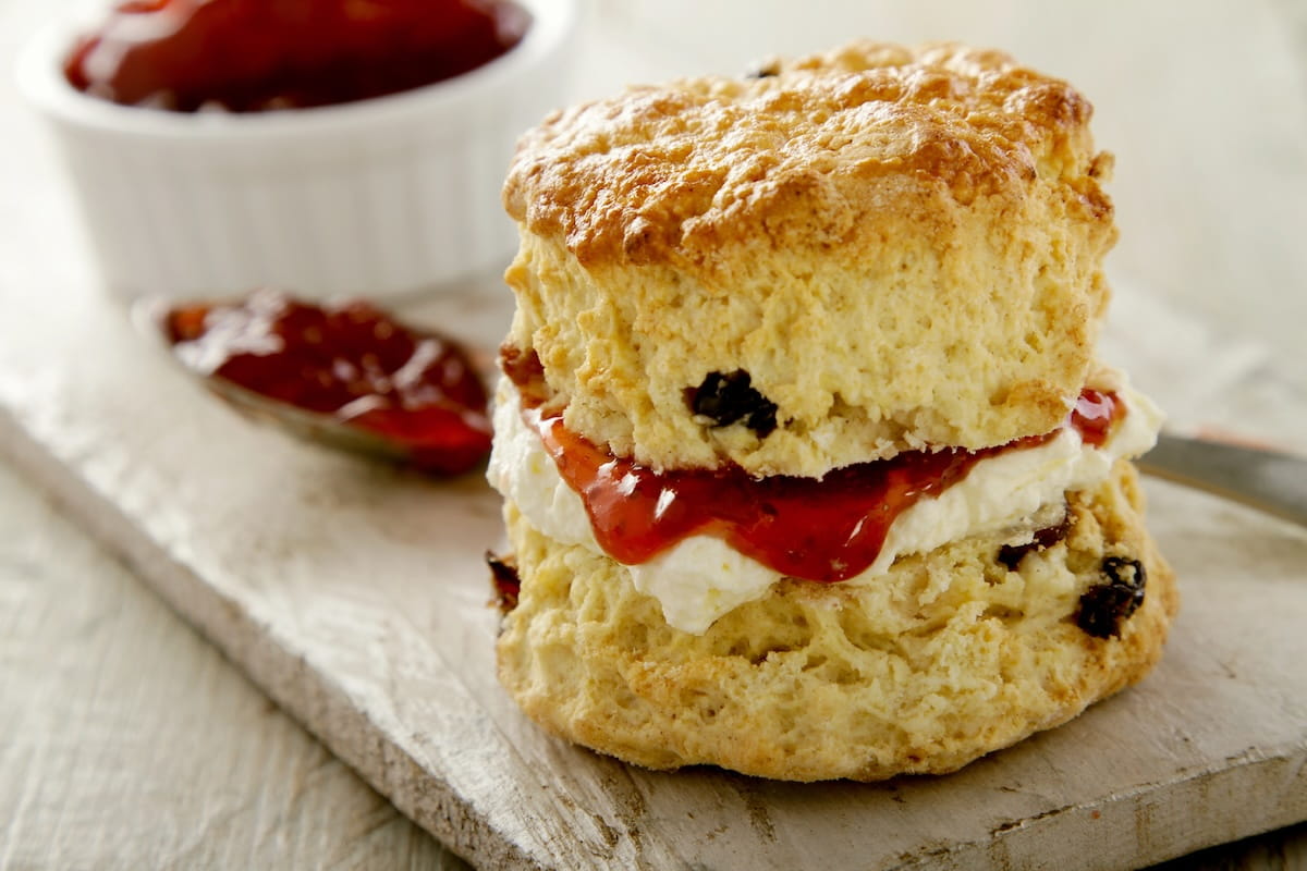 A close-up shot of a traditional cream tea presentation, featuring a freshly baked fruit scone split in half and layered with thick clotted cream and glossy red strawberry jam. The golden-brown scone sits on a rustic wooden serving board, with a small white bowl of extra jam and a spoon visible in the softly blurred background.