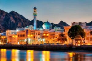 Evening view of Mutrah Corniche in Muscat, Oman, featuring a mosque with a blue-tiled dome and minaret illuminated by warm lights. The waterfront reflects the city lights, with traditional white buildings and rugged mountains in the background under a clear sky.