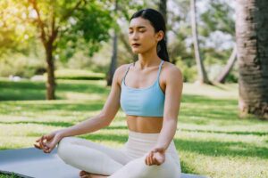 woman meditating in a peaceful park setting, sitting cross-legged on green grass with trees in the background on a sunny day
