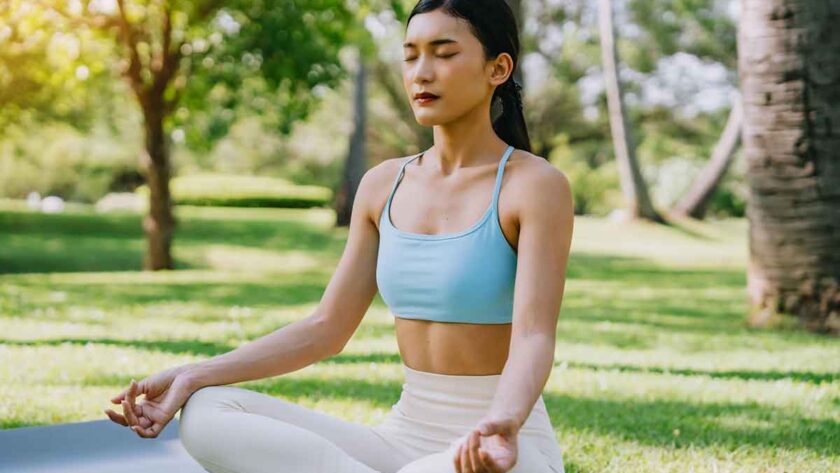 woman meditating in a peaceful park setting, sitting cross-legged on green grass with trees in the background on a sunny day