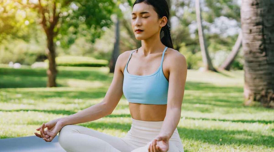 woman meditating in a peaceful park setting, sitting cross-legged on green grass with trees in the background on a sunny day