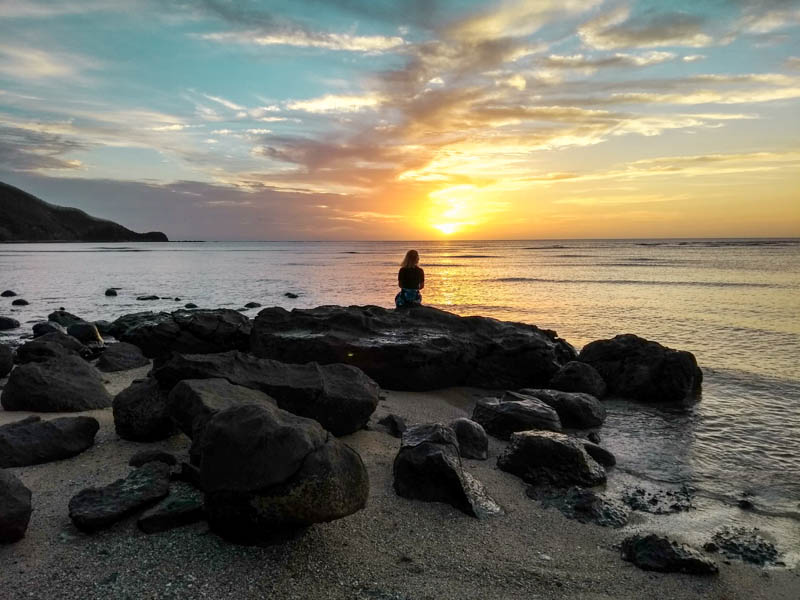 A silhouette of a girl sitting on rocks watching the sun set over the sea in Fiji. 