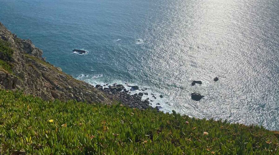 Sunlit Atlantic Ocean from Portuguese coastal cliff. Silver light reflects on water surface around dark rocky outcrops.