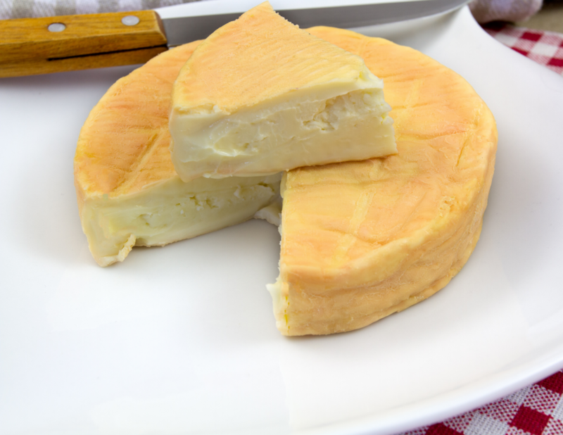 A wheel of Munster Cheese on a plate with a pie shaped piece cut out and resting on top of the wheel