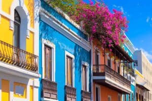 Vibrant, colorful buildings with ornate balconies line a narrow street in San Juan, Puerto Rico, against a backdrop of a clear blue sky. The bright orange and teal facades are adorned with flowering plants, reflecting the city's lively culture and architecture.
