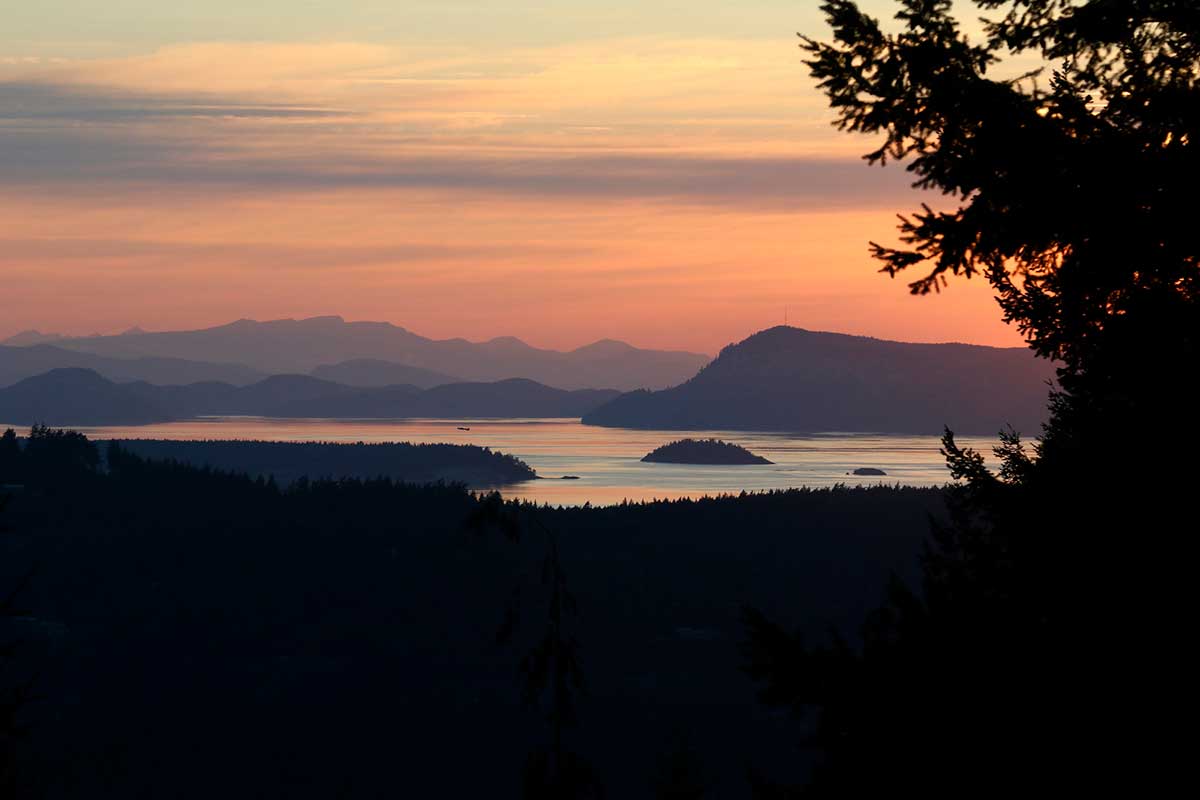 Stunning sunset view over the water from a ferry at Eastsound, Orcas Island, Washington. The sun dips below the horizon, casting a golden reflection on the calm waters, with clouds adding dramatic contrast to the sky.