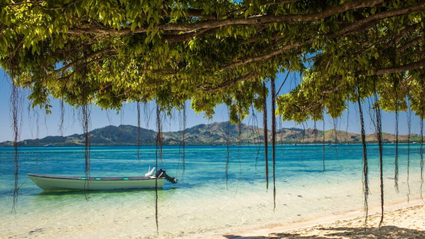 A quiet, sun-drenched beach in Fiji featuring a traditional-style boat floating in shallow transparent water under the shade of tropical foliage.