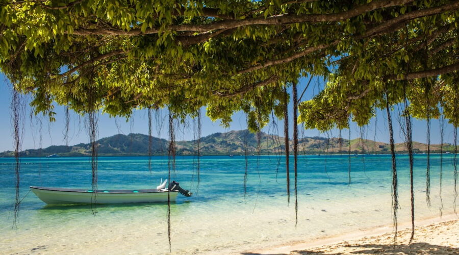 A quiet, sun-drenched beach in Fiji featuring a traditional-style boat floating in shallow transparent water under the shade of tropical foliage.