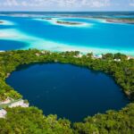 Aerial view of Bacalar, Mexico, showcasing the deep blue Cenote Azul surrounded by lush greenery, with the vibrant turquoise waters of the Lagoon of Seven Colors in the background.