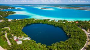 Aerial view of Bacalar, Mexico, showcasing the deep blue Cenote Azul surrounded by lush greenery, with the vibrant turquoise waters of the Lagoon of Seven Colors in the background.