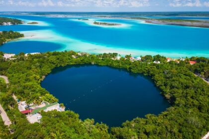 Aerial view of Bacalar, Mexico, showcasing the deep blue Cenote Azul surrounded by lush greenery, with the vibrant turquoise waters of the Lagoon of Seven Colors in the background.