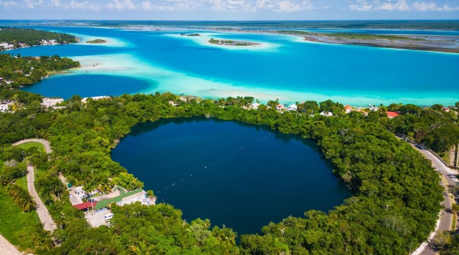 Aerial view of Bacalar, Mexico, showcasing the deep blue Cenote Azul surrounded by lush greenery, with the vibrant turquoise waters of the Lagoon of Seven Colors in the background.