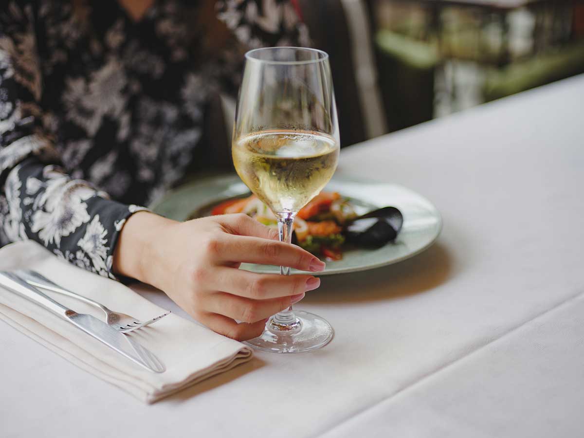 Close up photo of a woman's hand holding a glass of white wine and a seafood dish on the table in the background.