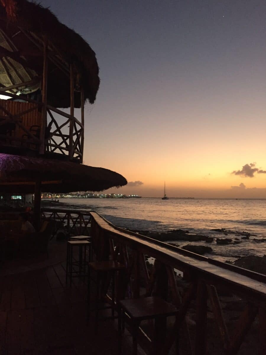 A peaceful beachfront restaurant in Cozumel at sunset, with thatched-roof palapas and a view of the ocean glowing under a golden sky.