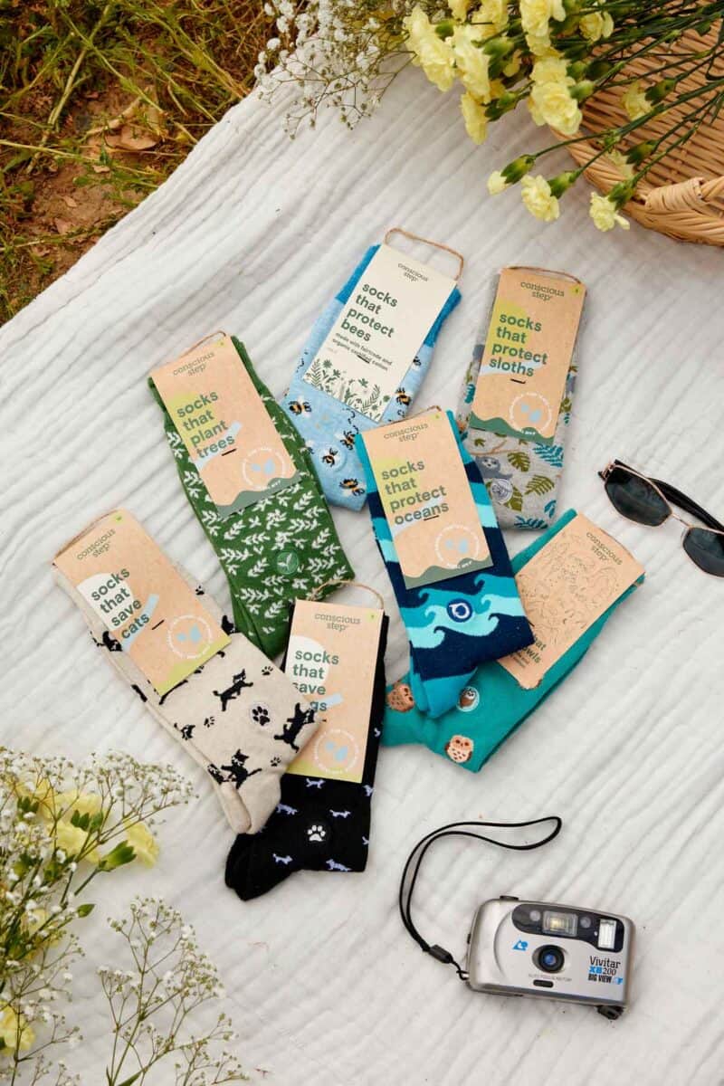 A flat lay of several pairs of colorful Conscious Step socks arranged on a white blanket outdoors. Each pair has a label with text such as 'socks that protect bees,' 'socks that plant trees,' 'socks that protect sloths,' 'socks that protect oceans,' 'socks that save cats,' and 'socks that save dogs.' The socks feature matching patterns like bees, trees, sloths, waves, and paw prints. Surrounding the socks are yellow flowers, a pair of sunglasses, a woven basket, and a silver film camera.