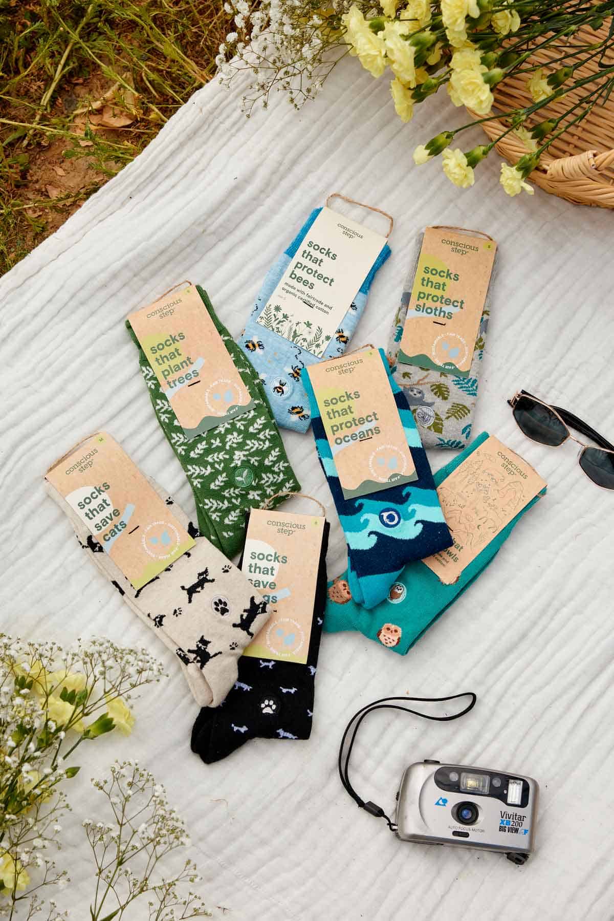 A flat lay of several pairs of colorful Conscious Step socks arranged on a white blanket outdoors. Each pair has a label with text such as 'socks that protect bees,' 'socks that plant trees,' 'socks that protect sloths,' 'socks that protect oceans,' 'socks that save cats,' and 'socks that save dogs.' The socks feature matching patterns like bees, trees, sloths, waves, and paw prints. Surrounding the socks are yellow flowers, a pair of sunglasses, a woven basket, and a silver film camera.