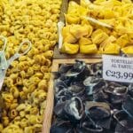 Close-up of a Bologna market stall displaying three varieties of fresh handmade pasta: small golden tortellini, larger yellow tortelloni, and black truffle-filled tortelloni labeled “Tortelloni al Tartufo €23,99,” with metal tongs resting on the tray.