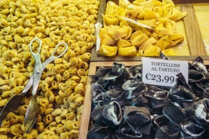 Close-up of a Bologna market stall displaying three varieties of fresh handmade pasta: small golden tortellini, larger yellow tortelloni, and black truffle-filled tortelloni labeled “Tortelloni al Tartufo €23,99,” with metal tongs resting on the tray.