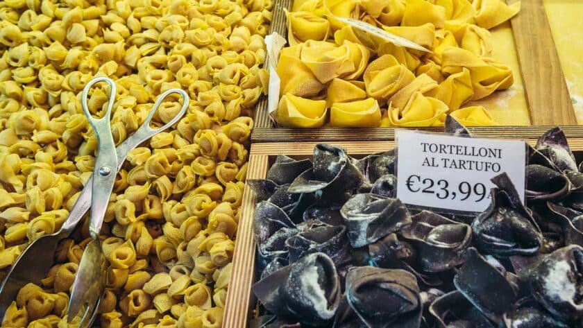 Close-up of a Bologna market stall displaying three varieties of fresh handmade pasta: small golden tortellini, larger yellow tortelloni, and black truffle-filled tortelloni labeled “Tortelloni al Tartufo €23,99,” with metal tongs resting on the tray.