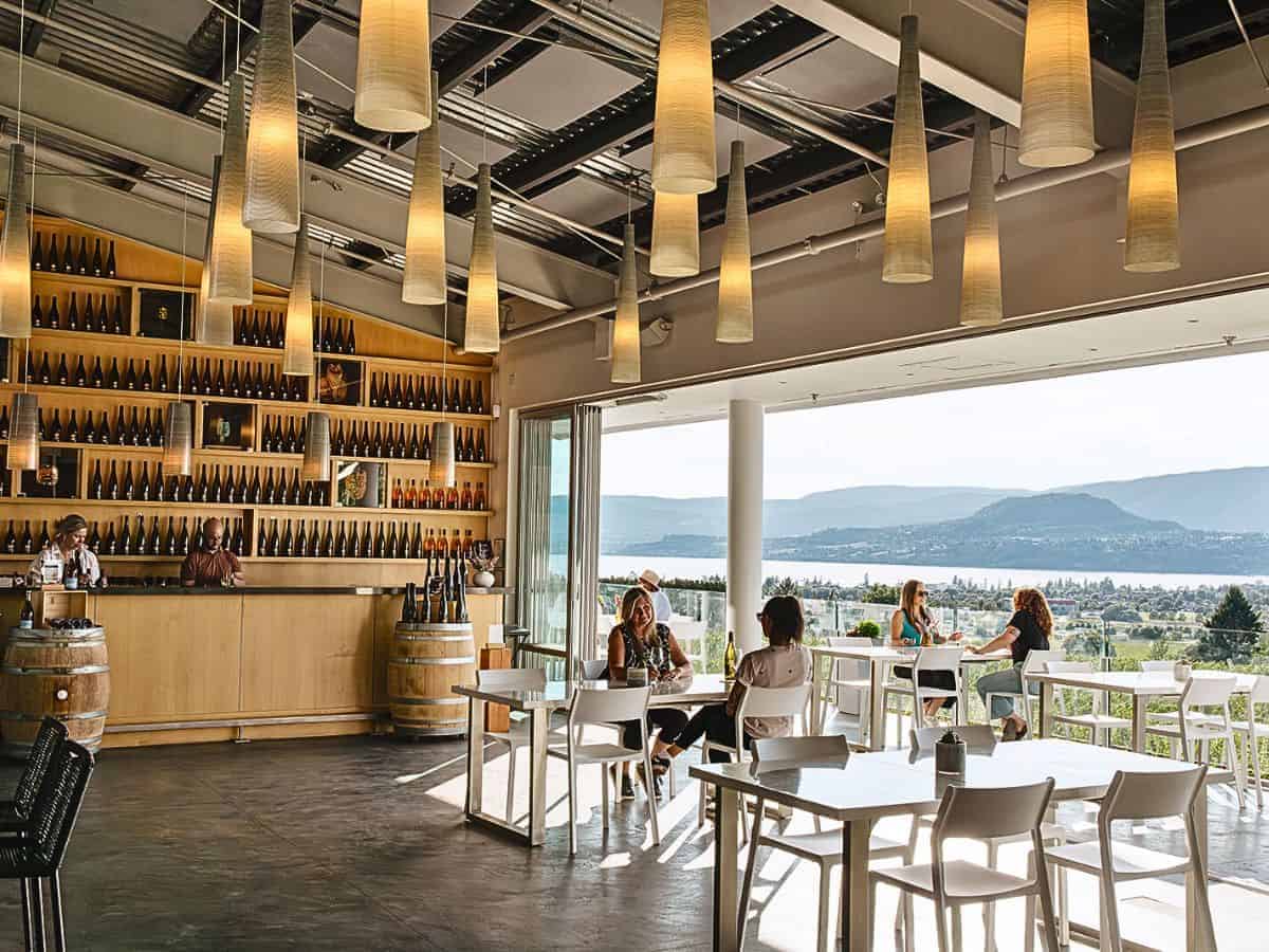 Tantalus Vineyards tasting room with bottle-lined wall, modern hanging lights, and balcony seating overlooking Lake Okanagan.