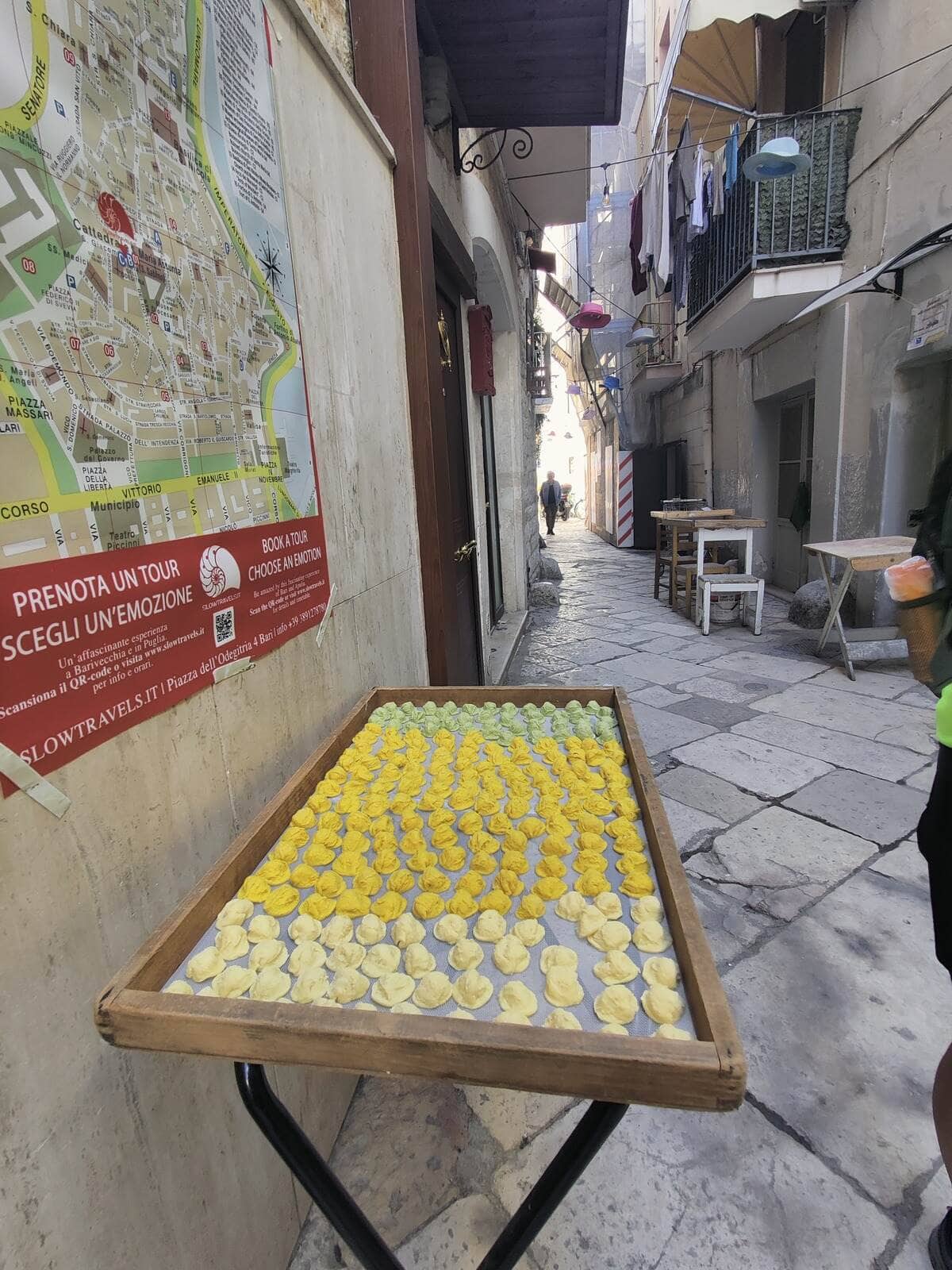 Colorful orecchiette drying on a wooden tray along a narrow stone street in Bari’s Old Town, with a city map on the wall and laundry hanging from balconies above.
