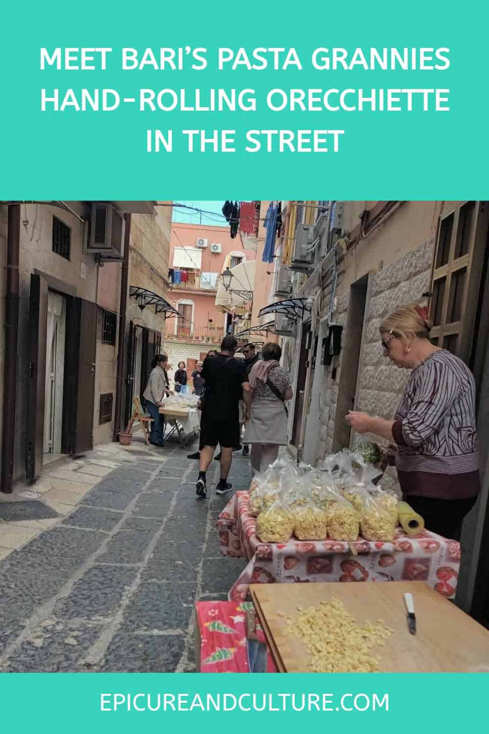 Pinterest pin that shows a photo of a street scene in Bari, Italy, where older women hand-roll orecchiette pasta at tables set up along a narrow stone alley. Bags of freshly made pasta are displayed on a table in the foreground, while people walk and chat among the historic buildings.
