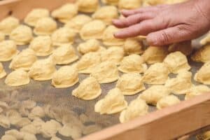 A close-up of freshly made orecchiette drying on a mesh tray, with an older woman’s hand gently arranging the pasta.