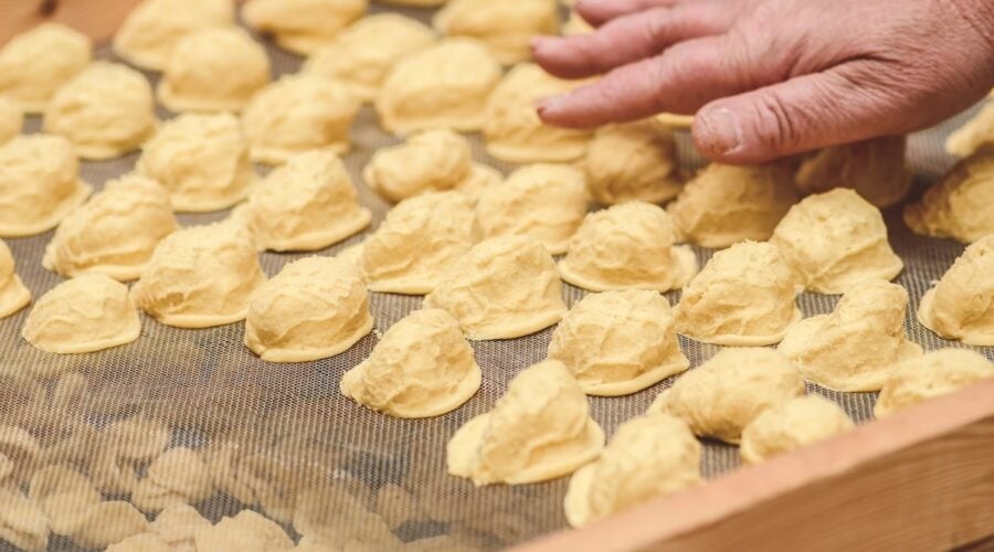 A close-up of freshly made orecchiette drying on a mesh tray, with an older woman’s hand gently arranging the pasta.